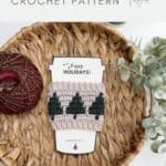 A crocheted mug cozy with green Christmas trees is displayed on a basket tray with yarn, greenery, and a "Happy Holidays" card. Text above reads "Christmas gift idea, Crochet Coffee Sleeve Pattern.