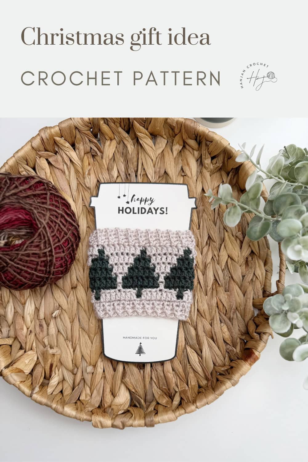 A crocheted mug cozy with green Christmas trees is displayed on a basket tray with yarn, greenery, and a "Happy Holidays" card. Text above reads "Christmas gift idea, Crochet Coffee Sleeve Pattern.