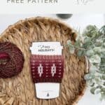 A red and white crocheted coffee sleeve, made from a festive Crochet Coffee Sleeve Pattern, is displayed on a woven tray next to a ball of yarn and green foliage.
