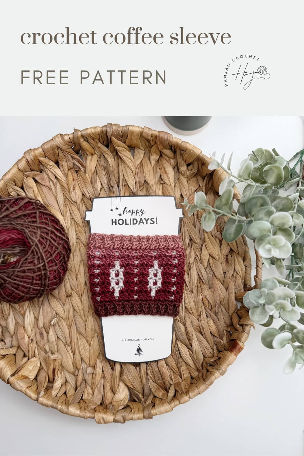 A red and white crocheted coffee sleeve, made from a festive Crochet Coffee Sleeve Pattern, is displayed on a woven tray next to a ball of yarn and green foliage.