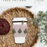 A reusable coffee cup with a Crochet Coffee Sleeve Pattern featuring green tree designs sits on a woven surface, next to a ball of yarn and some greenery.