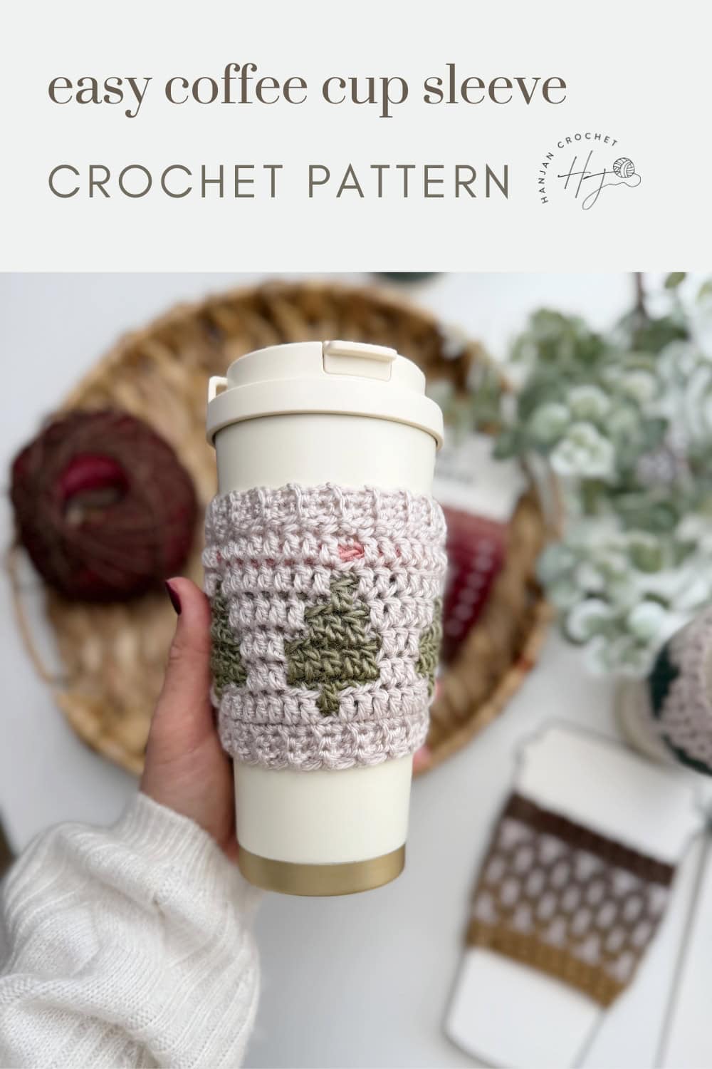 A hand holds a reusable cup with a crocheted sleeve featuring a green tree pattern. Yarn, crochet hook, and another Crochet Coffee Sleeve Pattern rest on a woven tray in the background.