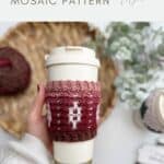 A hand holds a reusable cup adorned with a red and brown crochet coffee sleeve featuring a mosaic pattern; yarn, another cup, and the Crochet Coffee Sleeve Pattern are in the background.
