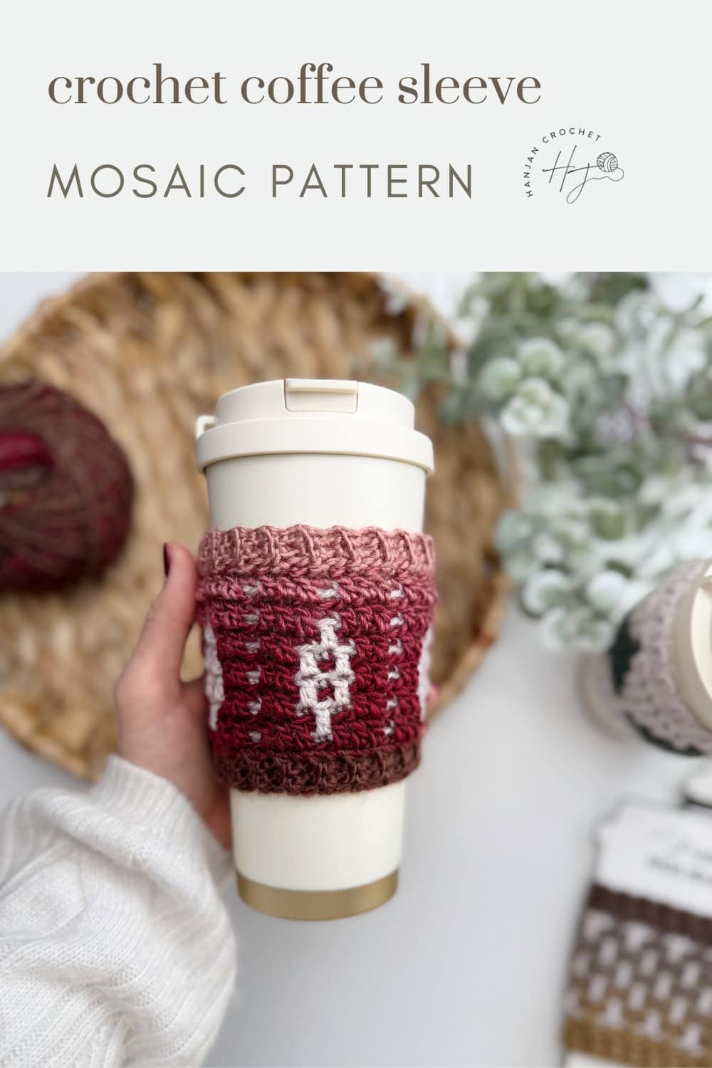 A hand holds a reusable cup adorned with a red and brown crochet coffee sleeve featuring a mosaic pattern; yarn, another cup, and the Crochet Coffee Sleeve Pattern are in the background.