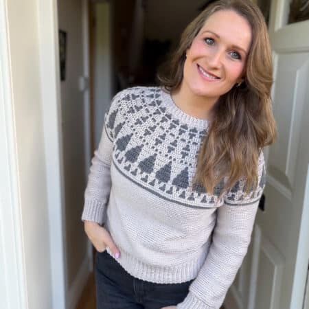 A woman with long brown hair, wearing a light crochet Christmas sweater with a patterned yoke and dark jeans, stands in a doorway, smiling at the camera.