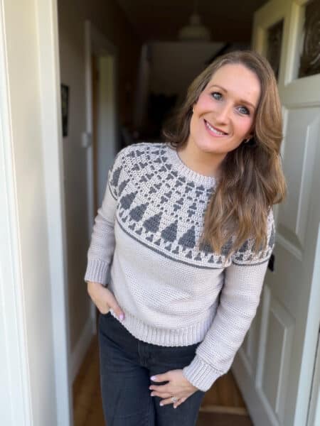A woman with long brown hair, wearing a light crochet Christmas sweater with a patterned yoke and dark jeans, stands in a doorway, smiling at the camera.