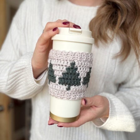 A person in a white sweater holds a reusable cup wrapped in a crochet coffee sleeve pattern, featuring charming green trees.