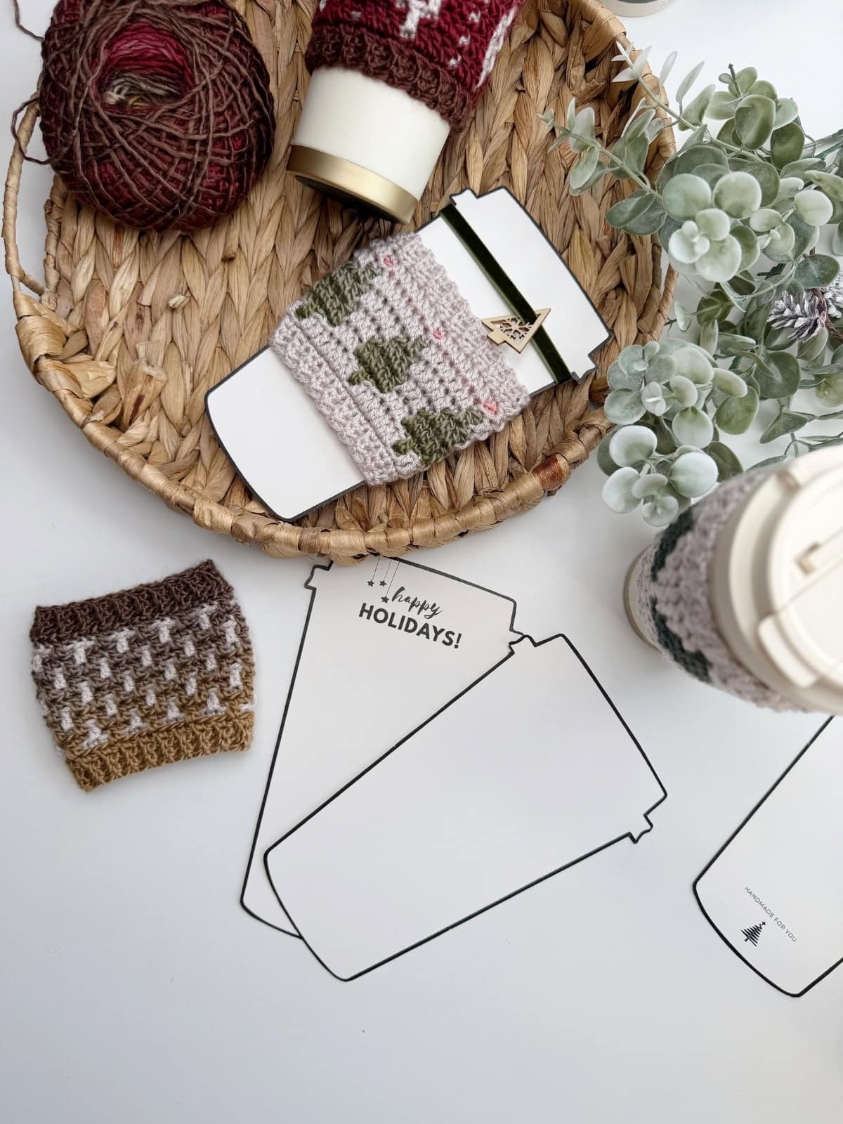 A wicker tray holds yarn, crochet cup sleeves, and a plant. Nearby, a to-go cup with a Crochet Coffee Sleeve Pattern and "Happy Holidays!" cards shaped like coffee cups sit on a white surface.
