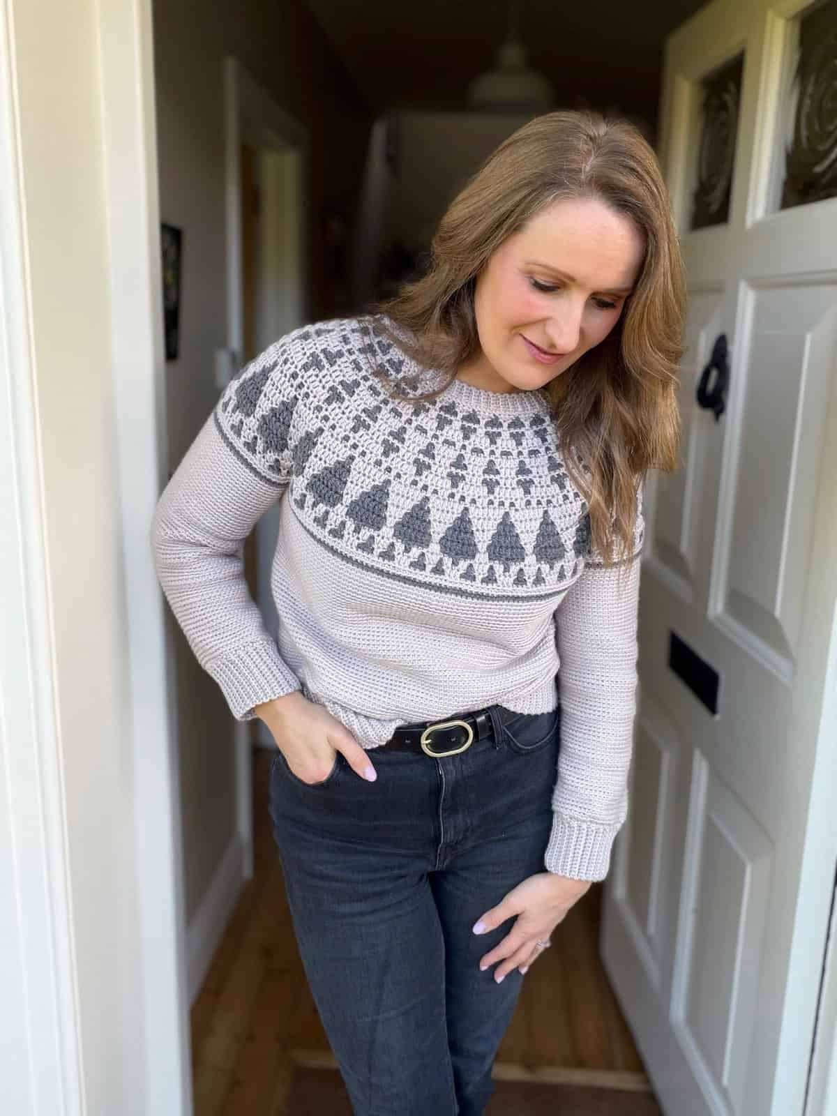 A woman stands in a doorway wearing a patterned knit sweater inspired by a crochet Christmas sweater pattern and black jeans, looking down with one hand in her pocket.