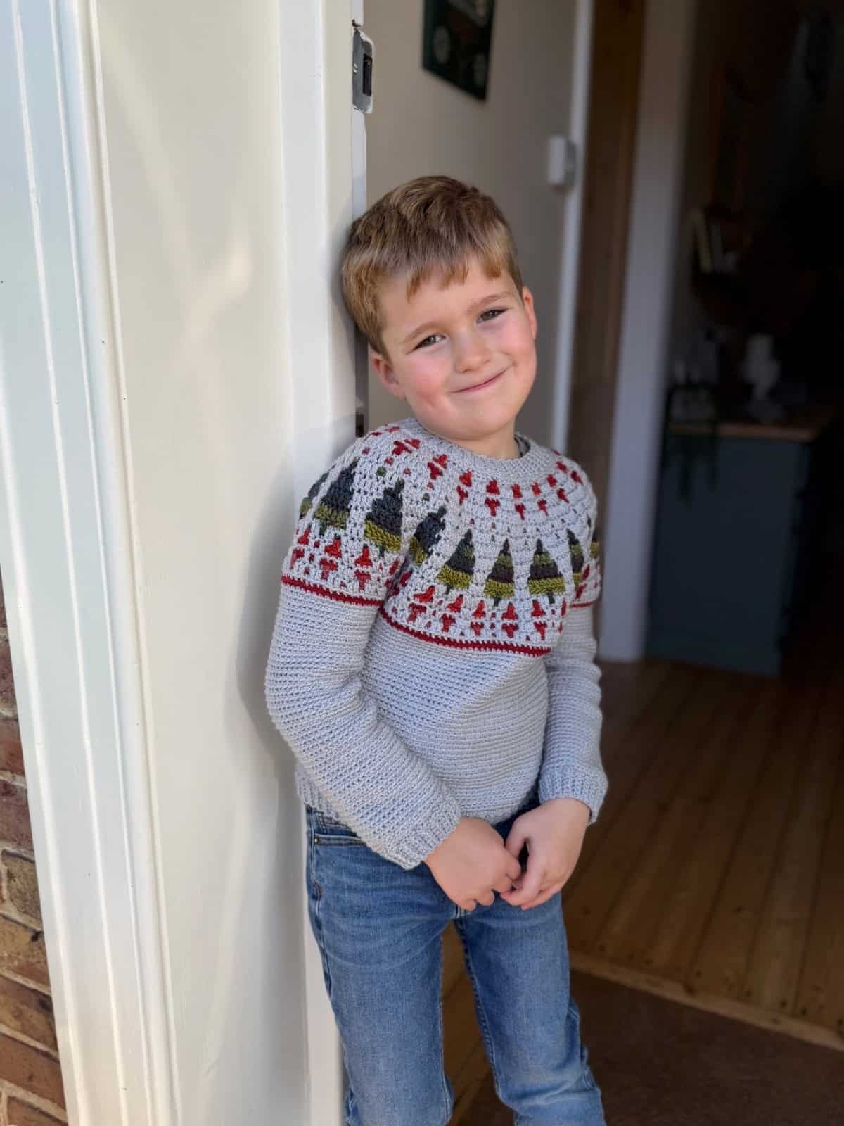 Young boy with light brown hair, wearing a crochet Christmas sweater and jeans, stands indoors leaning against a white doorframe, smiling at the camera.