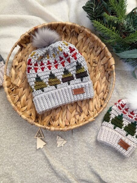 A woven basket holds a crochet winter hat with holiday patterns and a pom-pom, next to a matching smaller hat inspired by a christmas tree crochet hat pattern, on fabric with pine branches in the corner.
