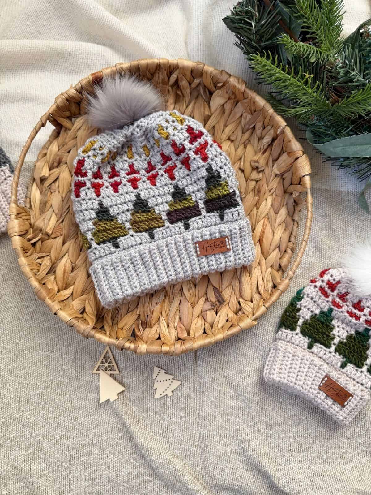 A woven basket holds a crochet winter hat with holiday patterns and a pom-pom, next to a matching smaller hat inspired by a christmas tree crochet hat pattern, on fabric with pine branches in the corner.