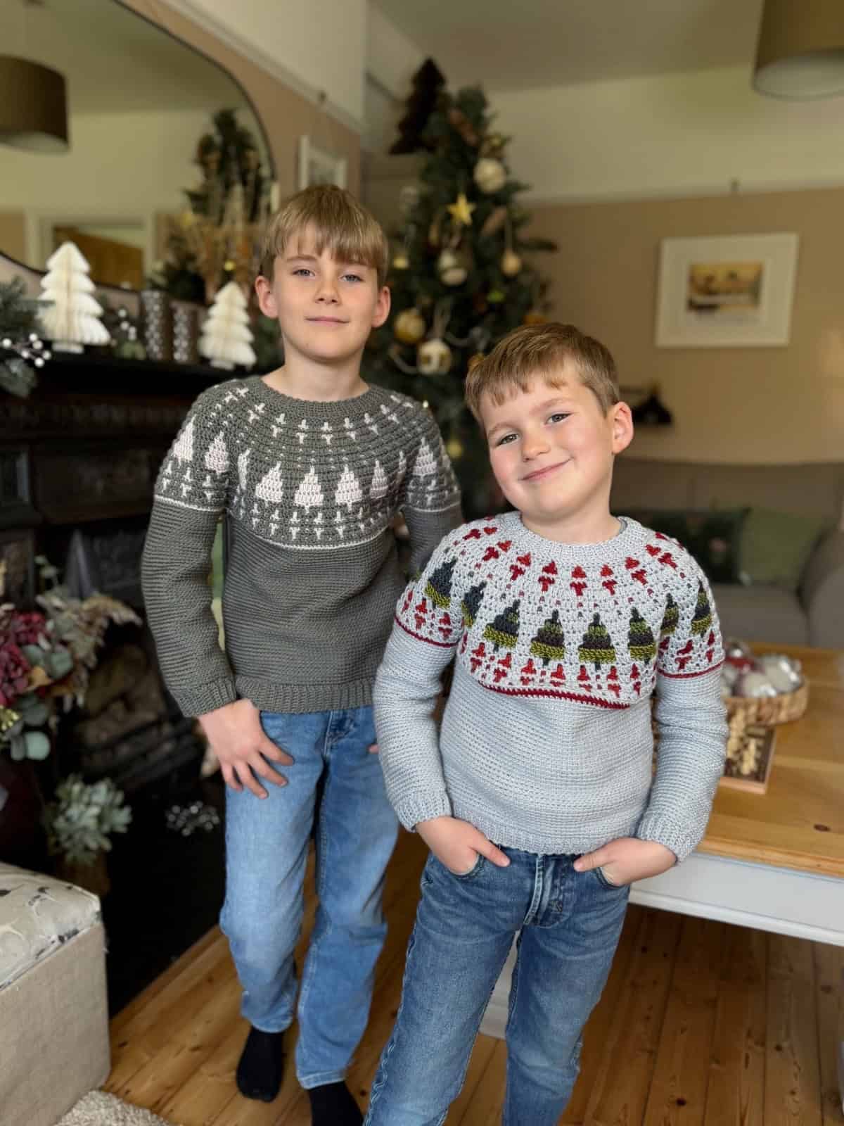Two boys in festive sweaters, featuring a cozy crochet Christmas sweater pattern, stand indoors near a decorated Christmas tree, smiling at the camera.