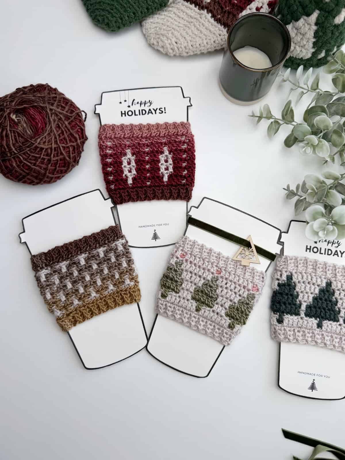 Four crochet cup cozies with holiday patterns displayed on cup-shaped cards, next to yarn, a candle, and eucalyptus leaves on a white surface—perfect for trying out a new Crochet Coffee Sleeve Pattern.