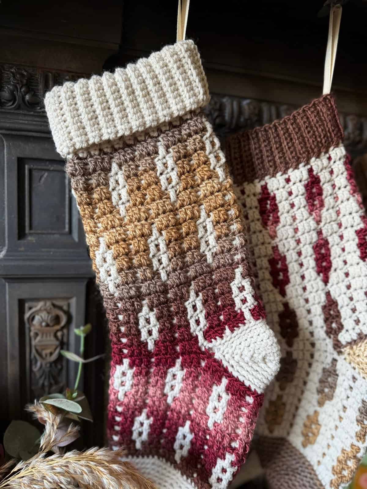 A crocheted Christmas stocking with a gradient pattern of cream, brown, and burgundy hangs by a fireplace, with another similar stocking partially visible beside it.