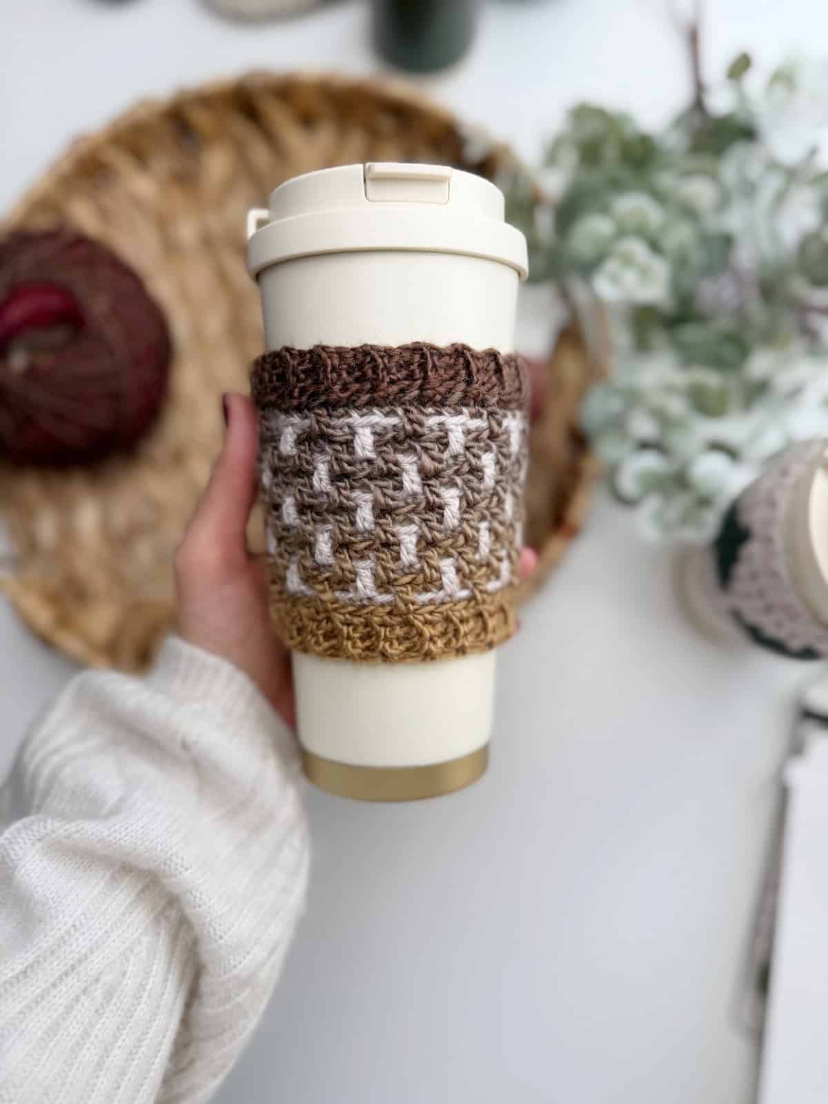 A hand holds a reusable coffee cup wrapped in a cozy crochet coffee sleeve pattern of brown, tan, and white; yarn and a plant add charm to the background.