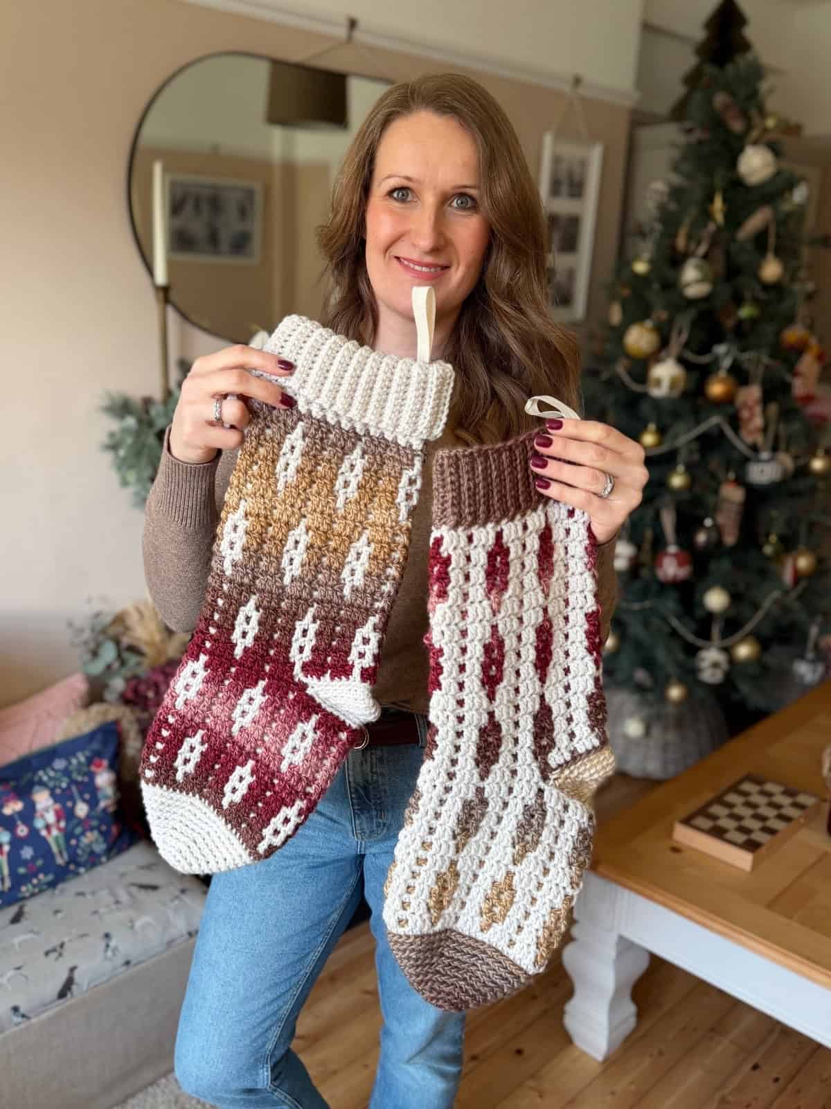 A woman stands in a living room holding two large, crochet Christmas stockings with patterns in brown, red, and cream colors. A decorated Christmas tree is visible in the background.