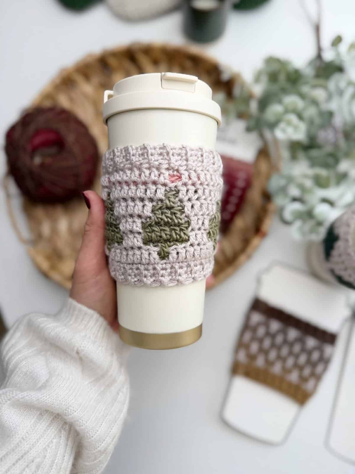 A hand holds a reusable cup with a crocheted sleeve; yarn, plants, and another cup sleeve are visible in the background, perfect inspiration for your next Crochet Coffee Sleeve Pattern.