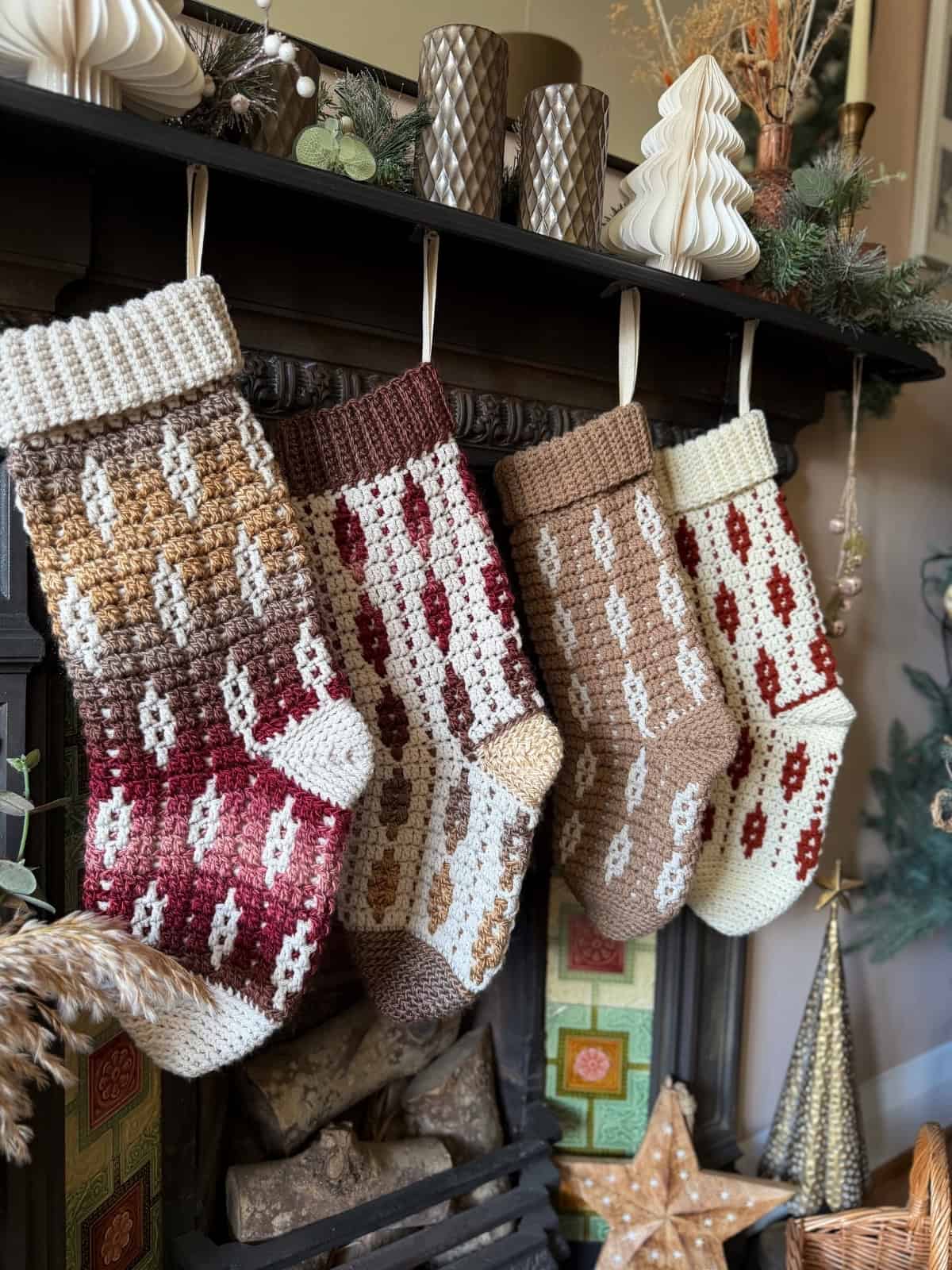 Four knitted Christmas stockings in earth tones hang from a decorated fireplace mantel with holiday ornaments and greenery.