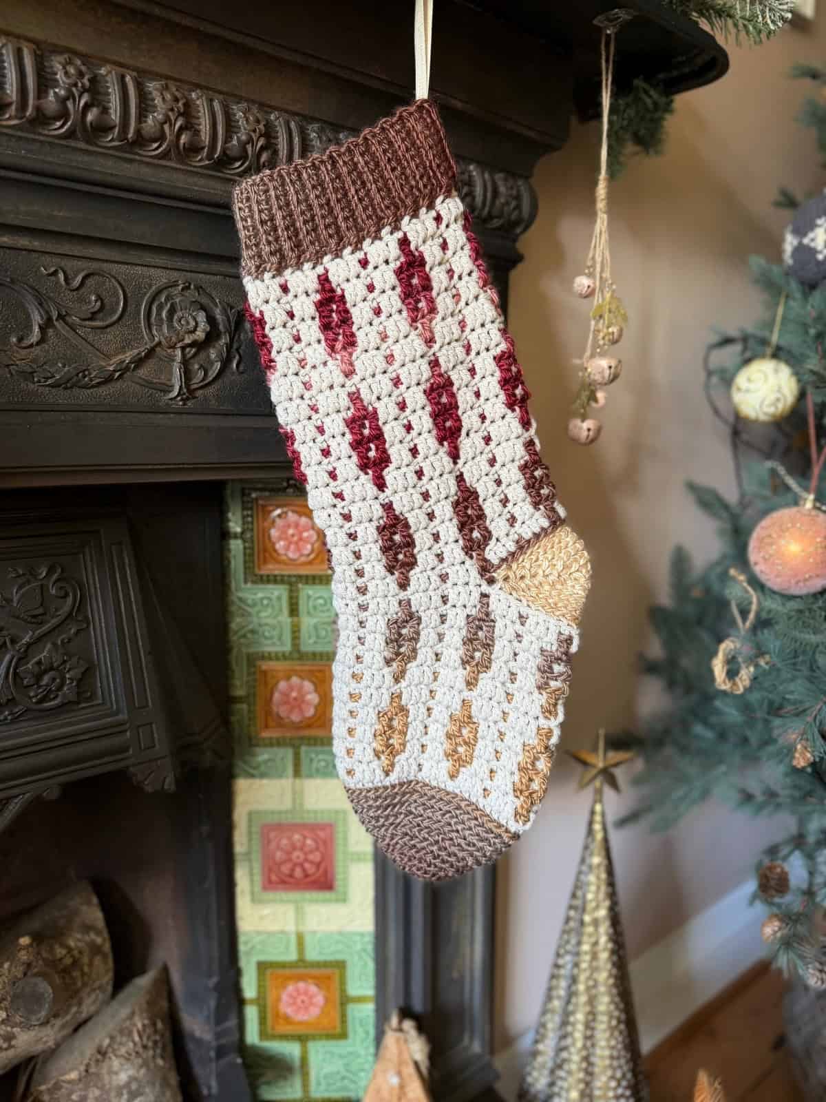 A crocheted Christmas stocking with a red, brown, and beige pattern hangs from a fireplace mantel next to a decorated tree.