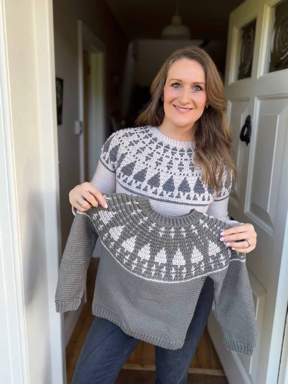 A woman stands in a doorway holding a child's sweater that matches her own, both featuring a patterned yoke design in grey and white—showcasing a beautiful crochet Christmas sweater pattern.
