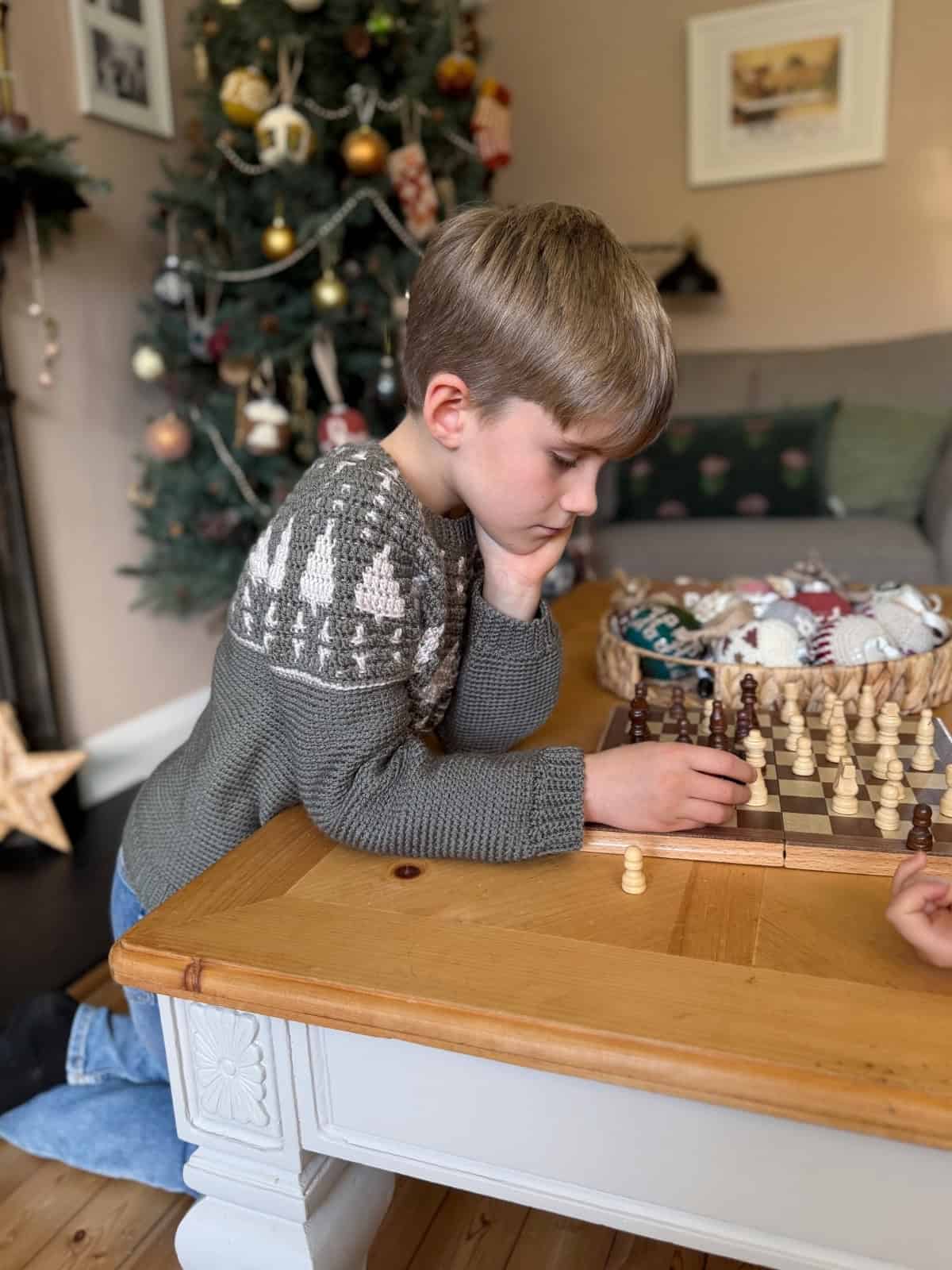 A young boy in a crochet Christmas sweater plays chess on a wooden table in a living room decorated for the holidays.