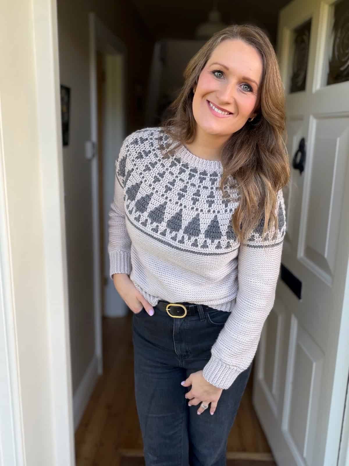 A woman with long brown hair, wearing a crochet Christmas sweater pattern and black jeans, stands at an open doorway, smiling at the camera.