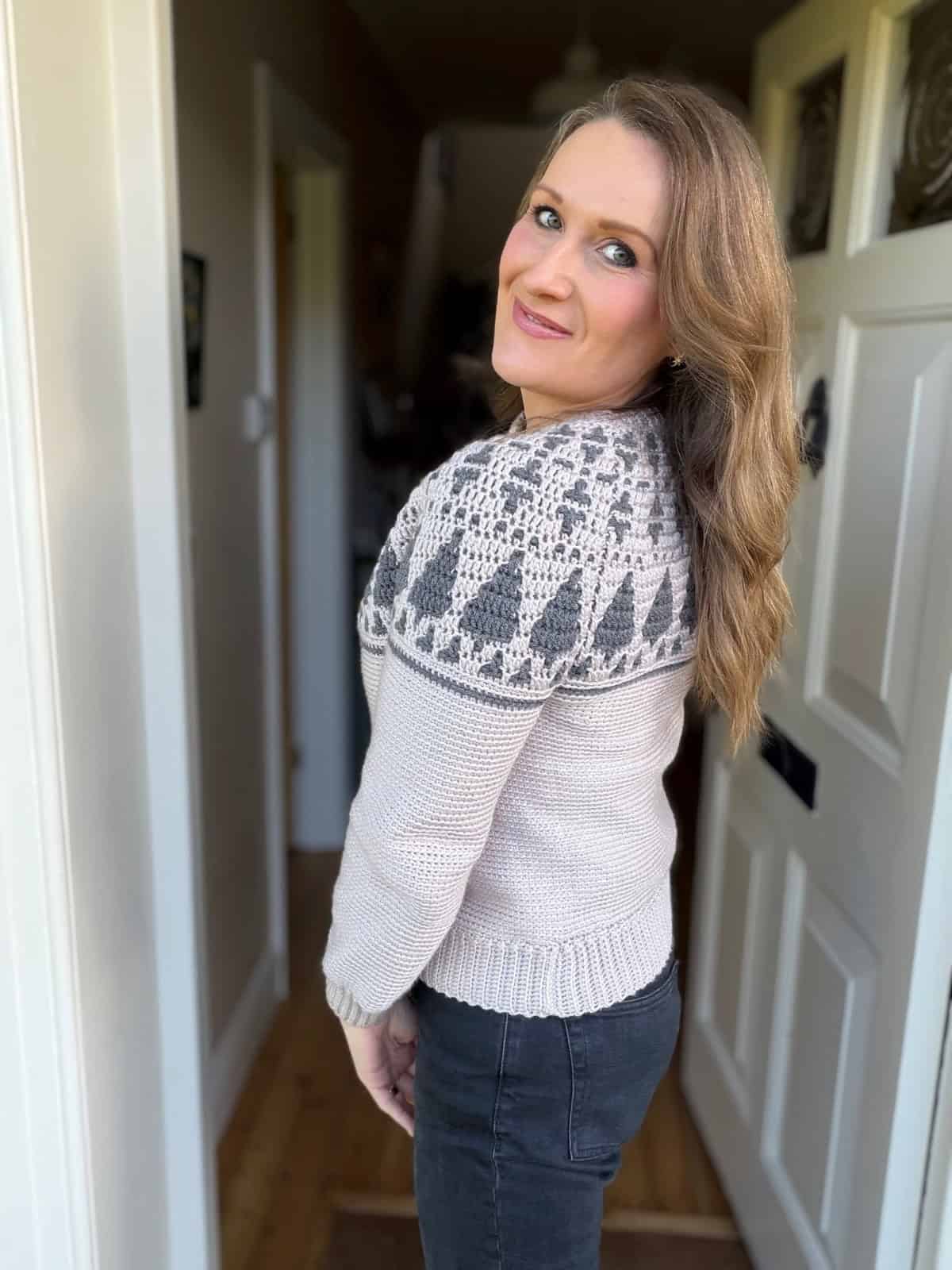 Woman with long, wavy hair stands in a doorway, wearing a light-colored crochet Christmas sweater pattern with a patterned yoke and dark jeans, looking back over her shoulder.