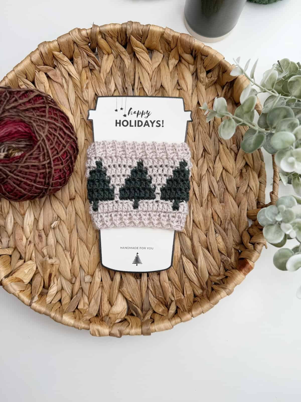 A crocheted cup cozy with green tree designs, made from a Crochet Coffee Sleeve Pattern, is displayed on a holiday-themed card and placed in a woven basket with yarn and greenery nearby.