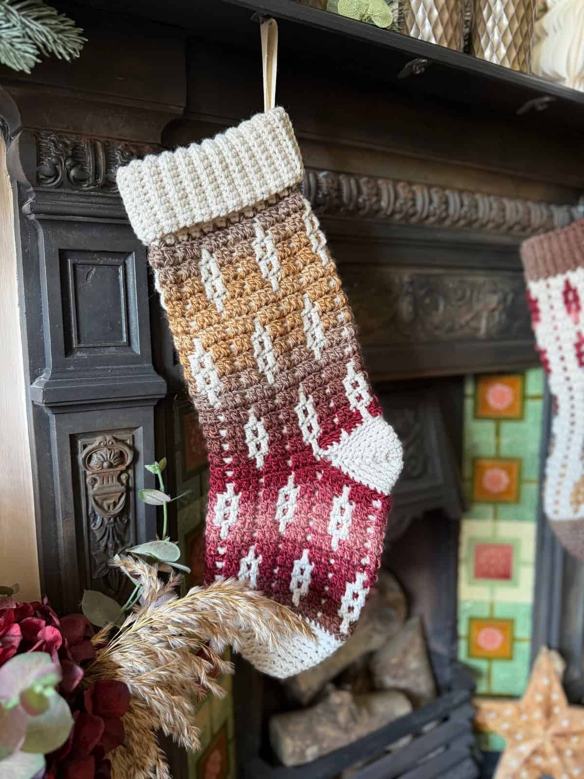 A crocheted Christmas stocking with red, brown, and cream colors hangs from a fireplace mantel decorated with plants and patterned tiles.