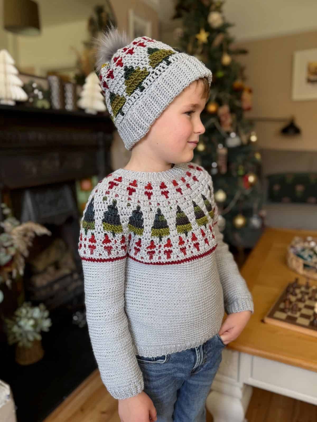 A young child stands indoors wearing a crochet sweater and a cozy christmas tree crochet hat pattern. A decorated Christmas tree and festive decorations are visible in the background.