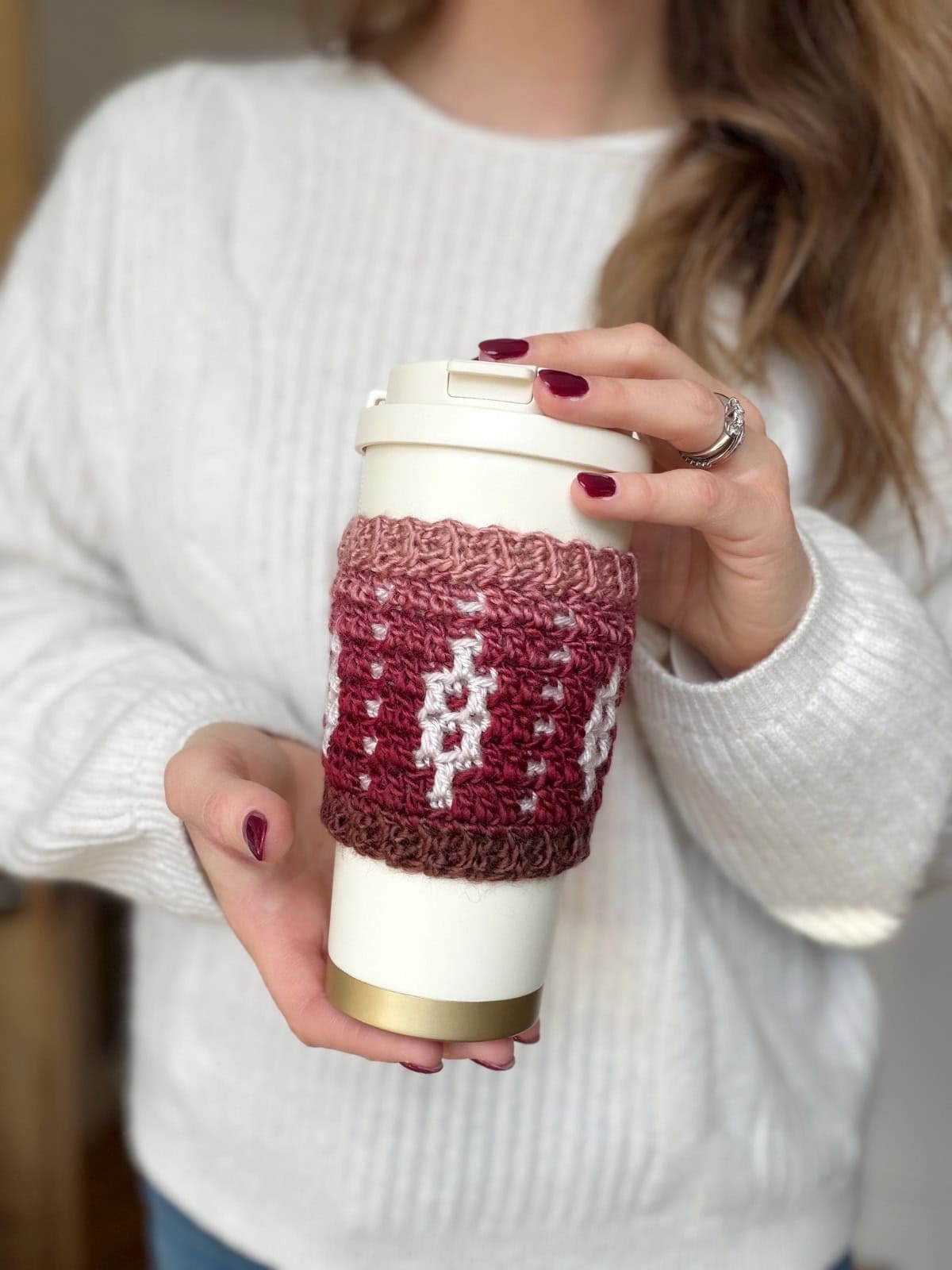 A person in a white sweater holds a reusable coffee cup with a maroon and pink crochet coffee sleeve pattern featuring a white design.