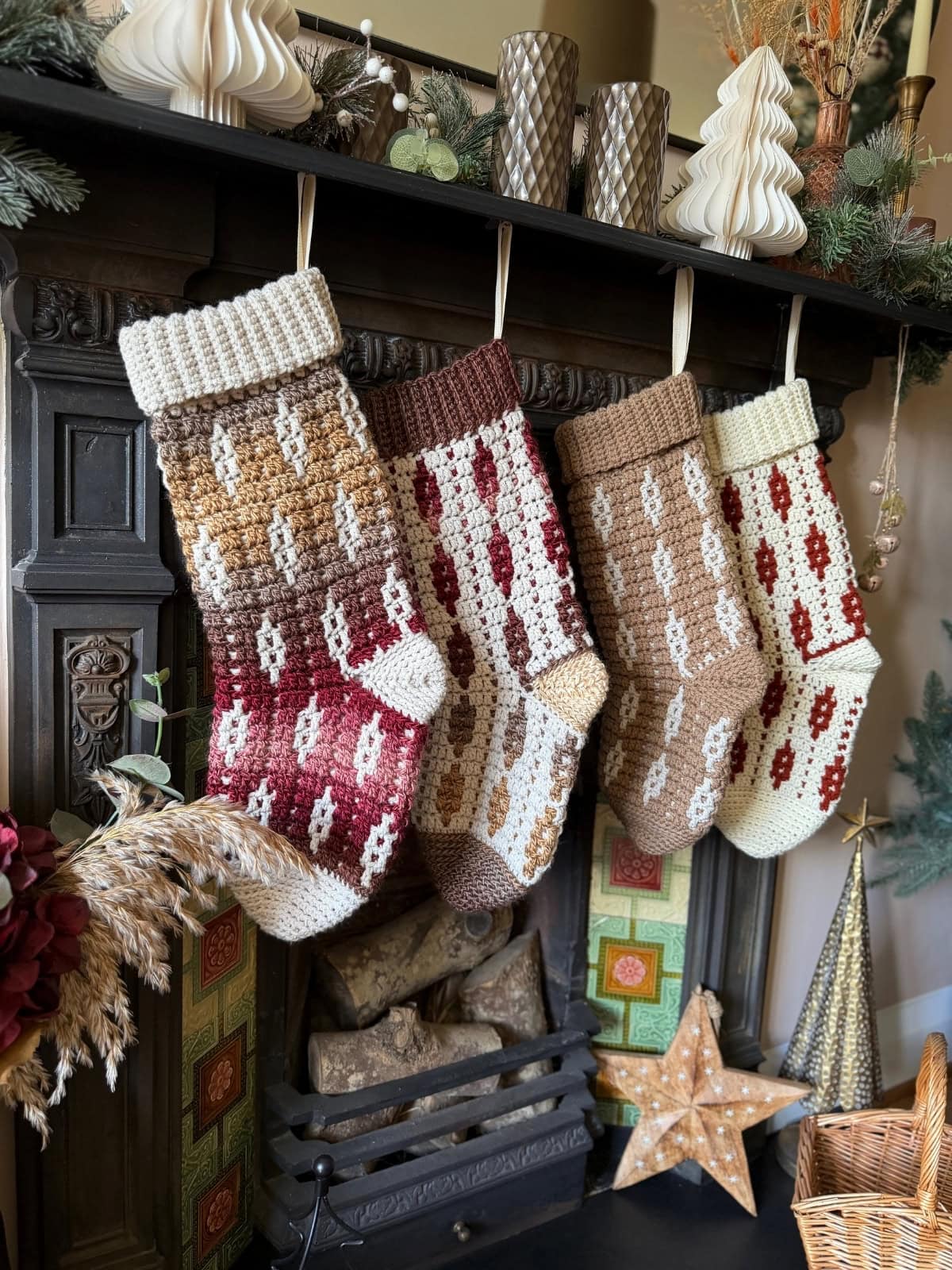 Four knitted Christmas stockings in earth tones hang from a fireplace mantel decorated with candles, paper trees, greenery, and festive ornaments.