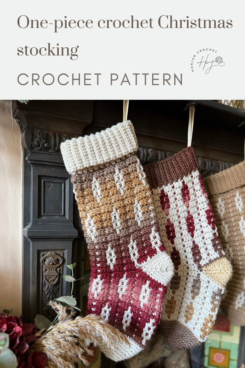 Two one-piece crochet Christmas stockings in red and brown tones hang from a dark wooden fireplace mantle, with decorative dried plants below—perfect inspiration if you’re learning how to crochet a Christmas stocking this holiday season.