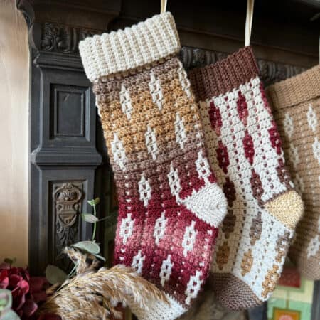 Two crocheted Christmas stockings with geometric patterns in shades of brown, cream, and red hang from a dark mantel above a fireplace.