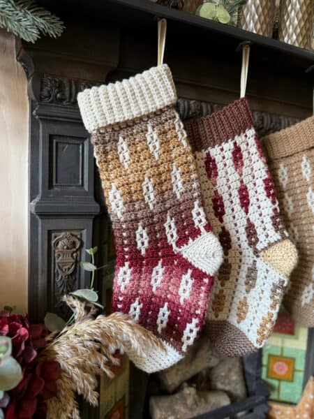 Two crocheted Christmas stockings with geometric patterns in shades of brown, cream, and red hang from a dark mantel above a fireplace.