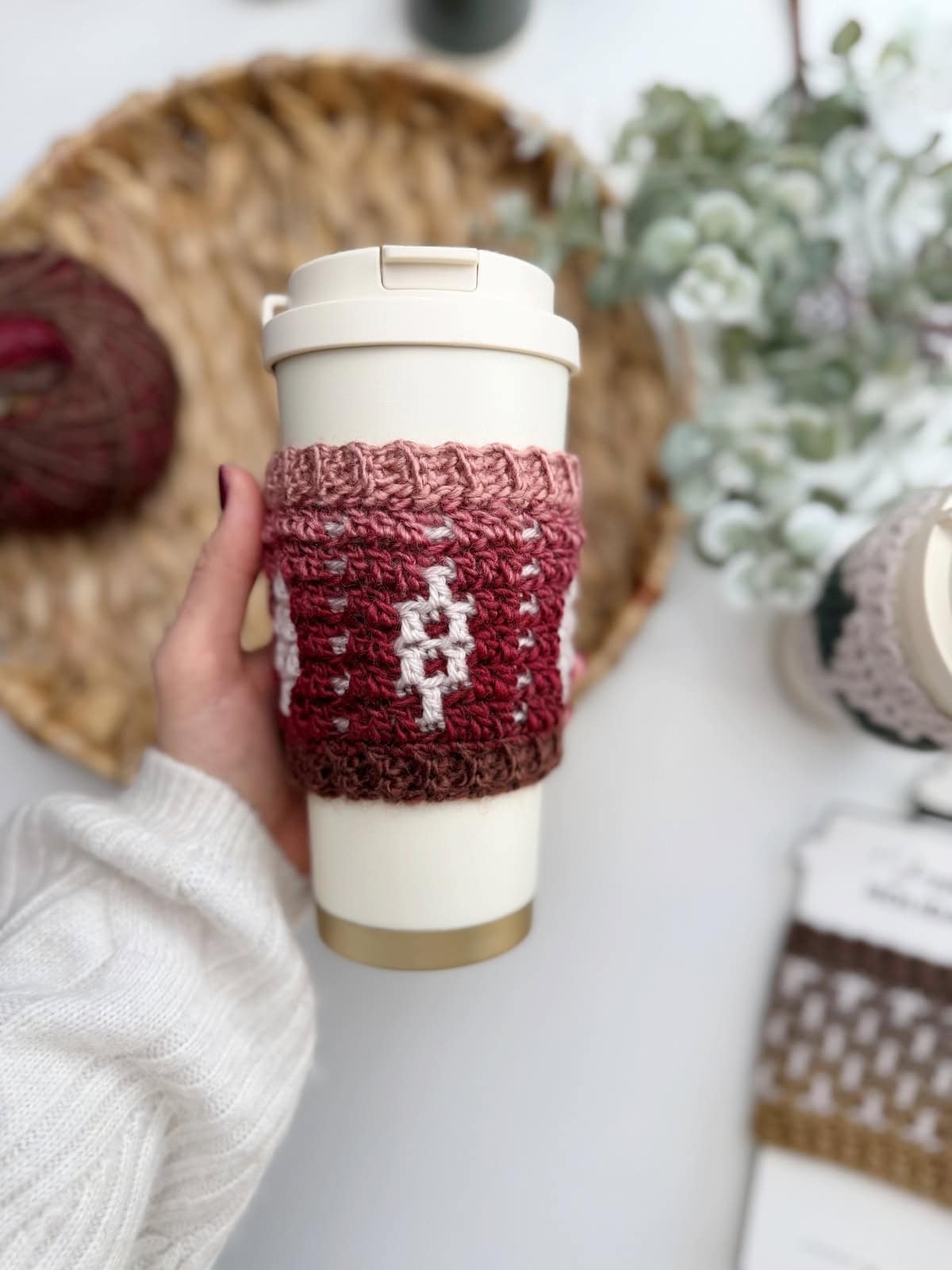 A hand holds a reusable travel cup wrapped in a pink and brown sleeve made from a Crochet Coffee Sleeve Pattern, set against a woven tray, yarn, and greenery.