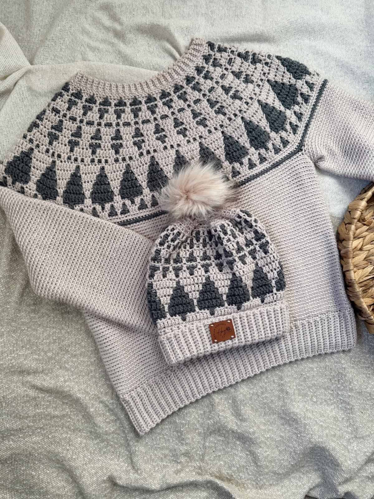 A knitted sweater and matching beanie with a geometric pattern in grey and cream, displayed on a light fabric surface. Perfect for winter, this set offers the cozy look of a crochet Christmas sweater pattern, with a faux fur pom-pom on the beanie.