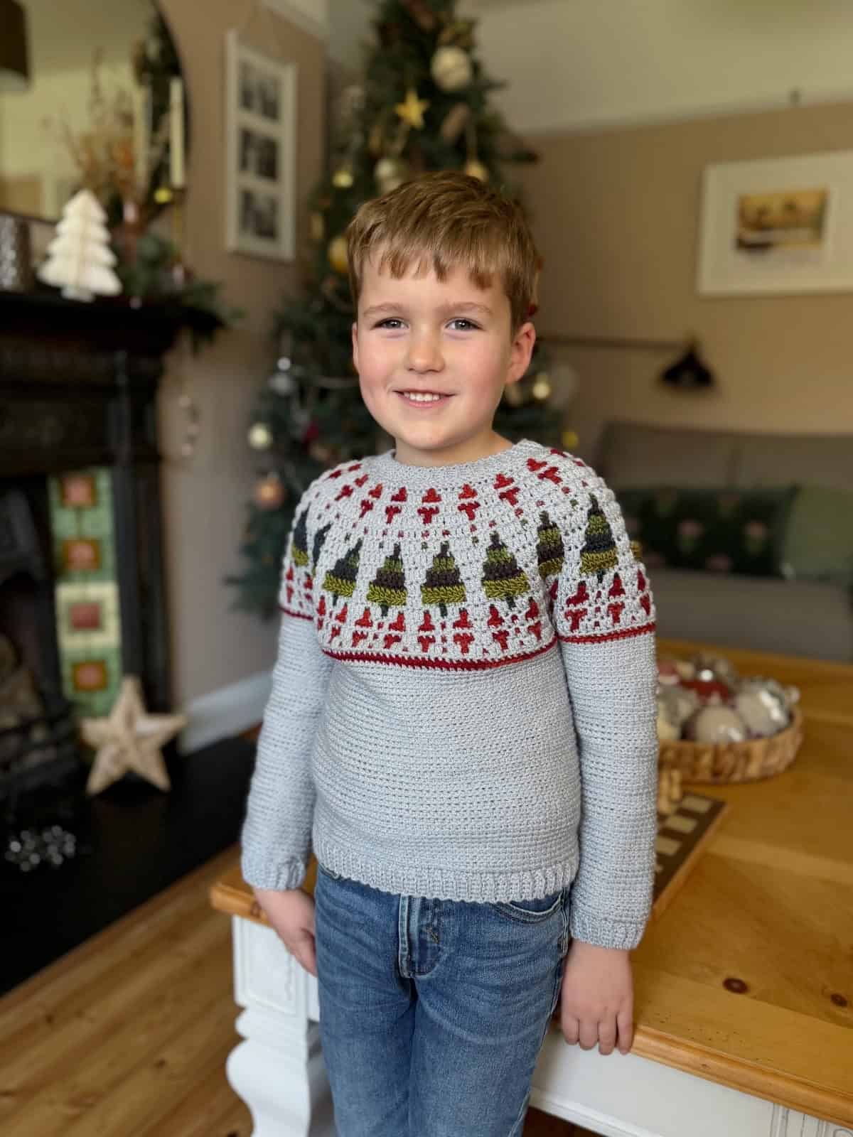 A young boy stands indoors smiling, wearing a gray crochet Christmas sweater with a pattern of Christmas trees and red accents. A decorated Christmas tree is visible in the background.