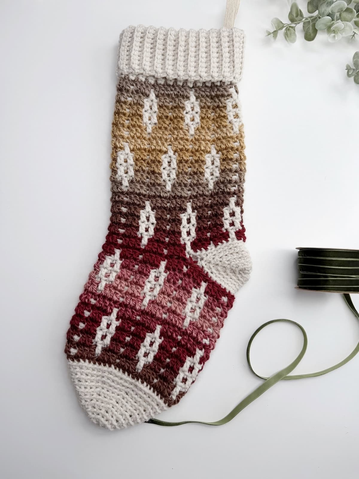A crocheted Christmas stocking with a gradient pattern in shades of beige, brown, and red, lying on a white surface next to a green ribbon and some greenery.