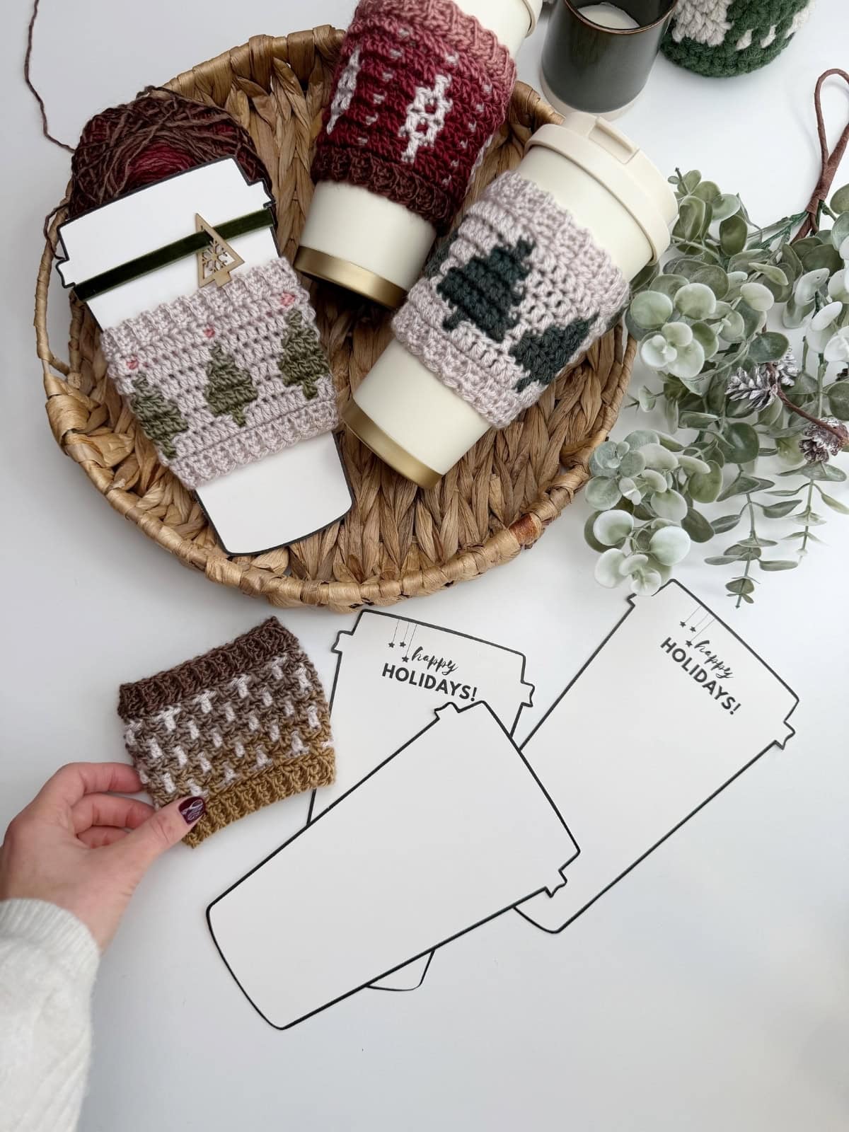 A hand holds a knitted cup sleeve near three reusable cups with holiday-themed sleeves on a wicker tray, next to paper cutouts shaped like coffee cups with "HAPPY HOLIDAYS!"—a festive scene perfect for showing off your Crochet Coffee Sleeve Pattern.
