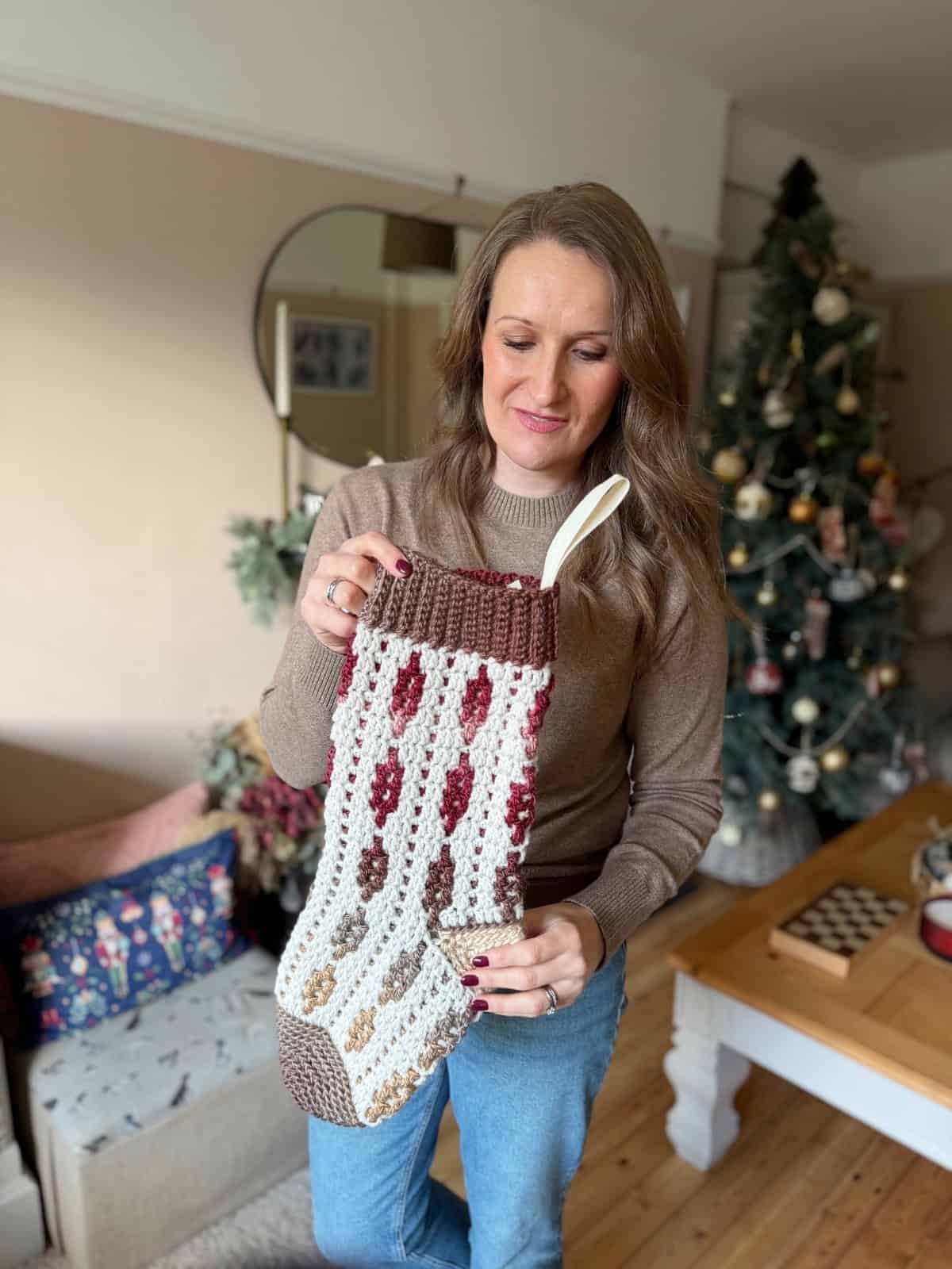 A woman standing in a living room holds a large crocheted Christmas stocking. A decorated Christmas tree and festive decor are visible in the background.