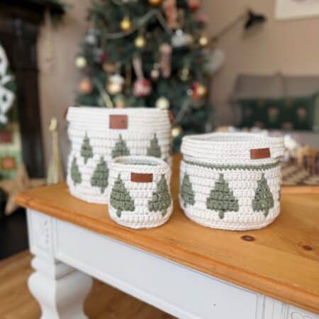 Three white christmas crochet baskets with green Christmas tree designs are displayed on a wooden table, with a decorated Christmas tree in the background.