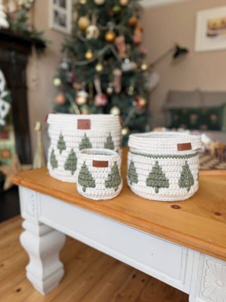 Three white christmas crochet baskets with green Christmas tree designs are displayed on a wooden table, with a decorated Christmas tree in the background.