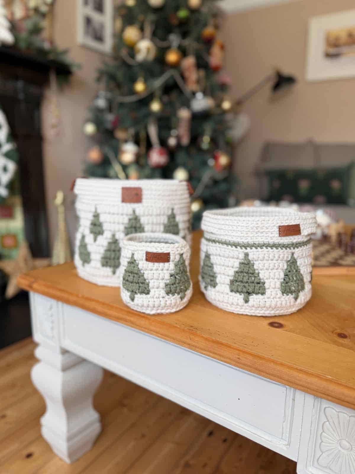 Three white christmas crochet baskets with green Christmas tree designs are displayed on a wooden table, with a decorated Christmas tree in the background.