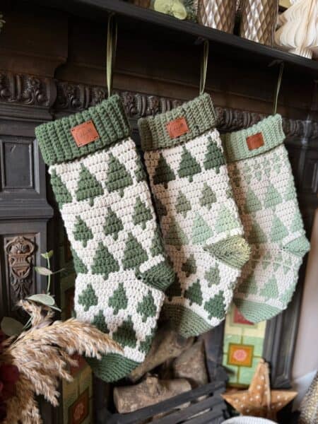 Three green and white Christmas stockings with tree patterns, crafted from a crochet stocking pattern, hang from hooks on a dark fireplace mantel.