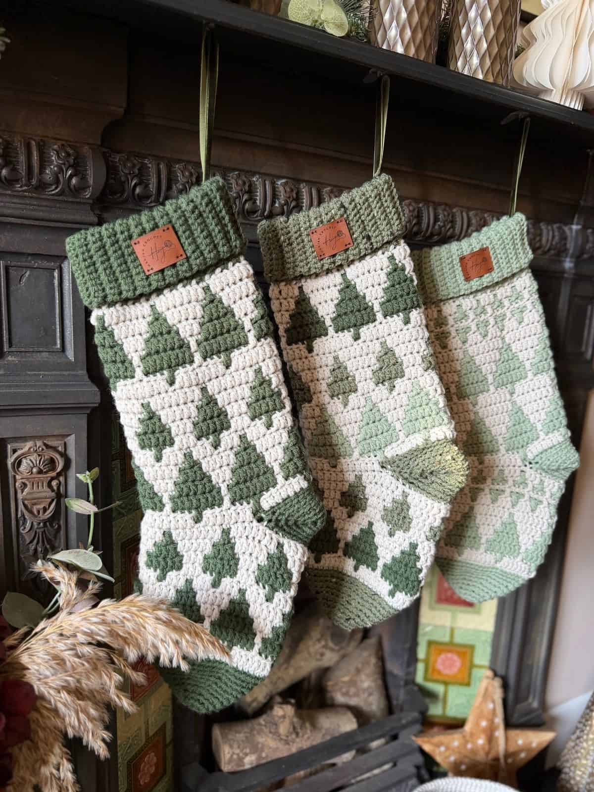 Three green and white Christmas stockings with tree patterns, crafted from a crochet stocking pattern, hang from hooks on a dark fireplace mantel.