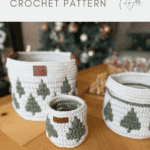 Three white christmas crochet baskets with green tree patterns are displayed on a wooden table, with a blurred Christmas tree and holiday decor in the background.