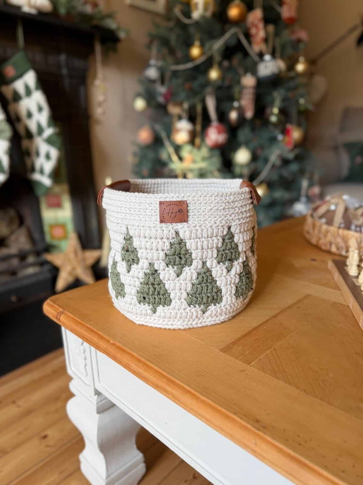 A christmas crochet basket with a green tree pattern and brown handles sits on a wooden coffee table in a decorated living room, with a Christmas tree glowing in the background.