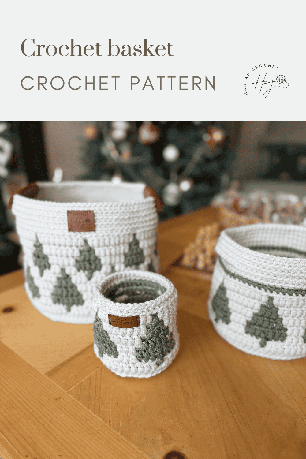 Three white christmas crochet baskets with green tree patterns are displayed on a wooden table, with a blurred Christmas tree and holiday decor in the background.